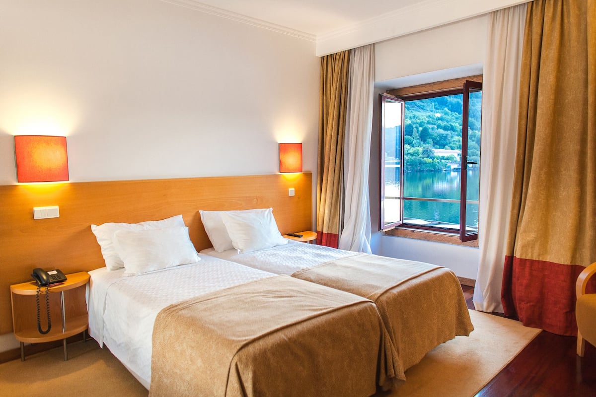 Habitación de hotel con dos camas, una mesita de noche con teléfono, una ventana con vista a un lago y una lámpara fijada a la pared.