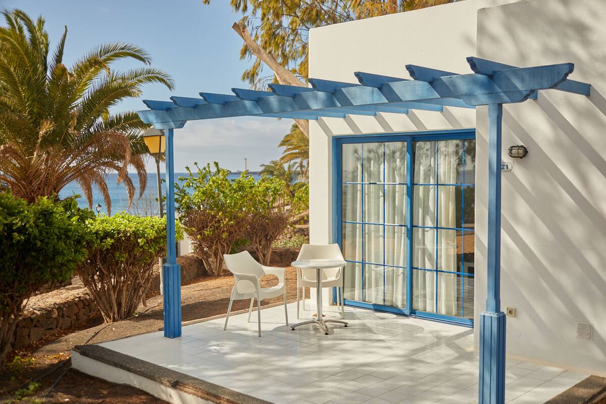 Un patio con dos sillas blancas y una pequeña mesa, una pérgola con detalles azules y vista al mar.