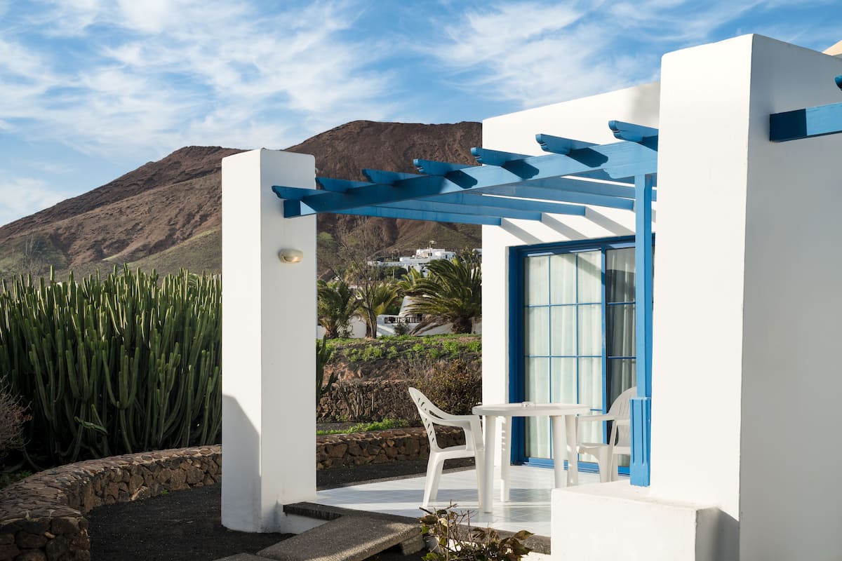 Un espacio exterior moderno con una mesa y sillas blancas, una pérgola azul y montañas al fondo.