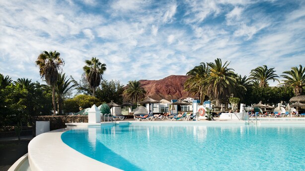 Une piscine entourée de chaises longues et de parasols, bordée de palmiers et avec une montagne en toile de fond.