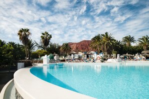 Une piscine entourée de chaises longues et de parasols, bordée de palmiers et avec une montagne en toile de fond.
