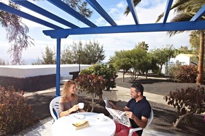 Dos personas sentadas en una mesa redonda bajo una pérgola azul, con vista a un jardín paisajístico y un edificio al fondo.