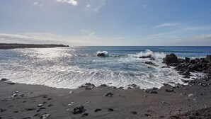 Une plage où les vagues se brisent sur le sable sombre et les rochers, sous un ciel dégagé.