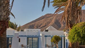 Un bâtiment blanc agrémenté de touches de bleu, une petite table et des chaises à l’extérieur, et une montagne en arrière-plan.