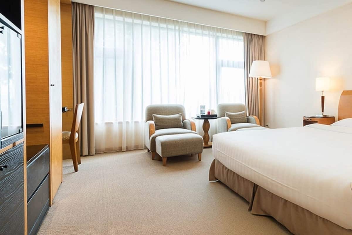 Quarto de hotel com uma cama grande, duas poltronas, uma mesinha e uma escrivaninha com abajur e telefone.