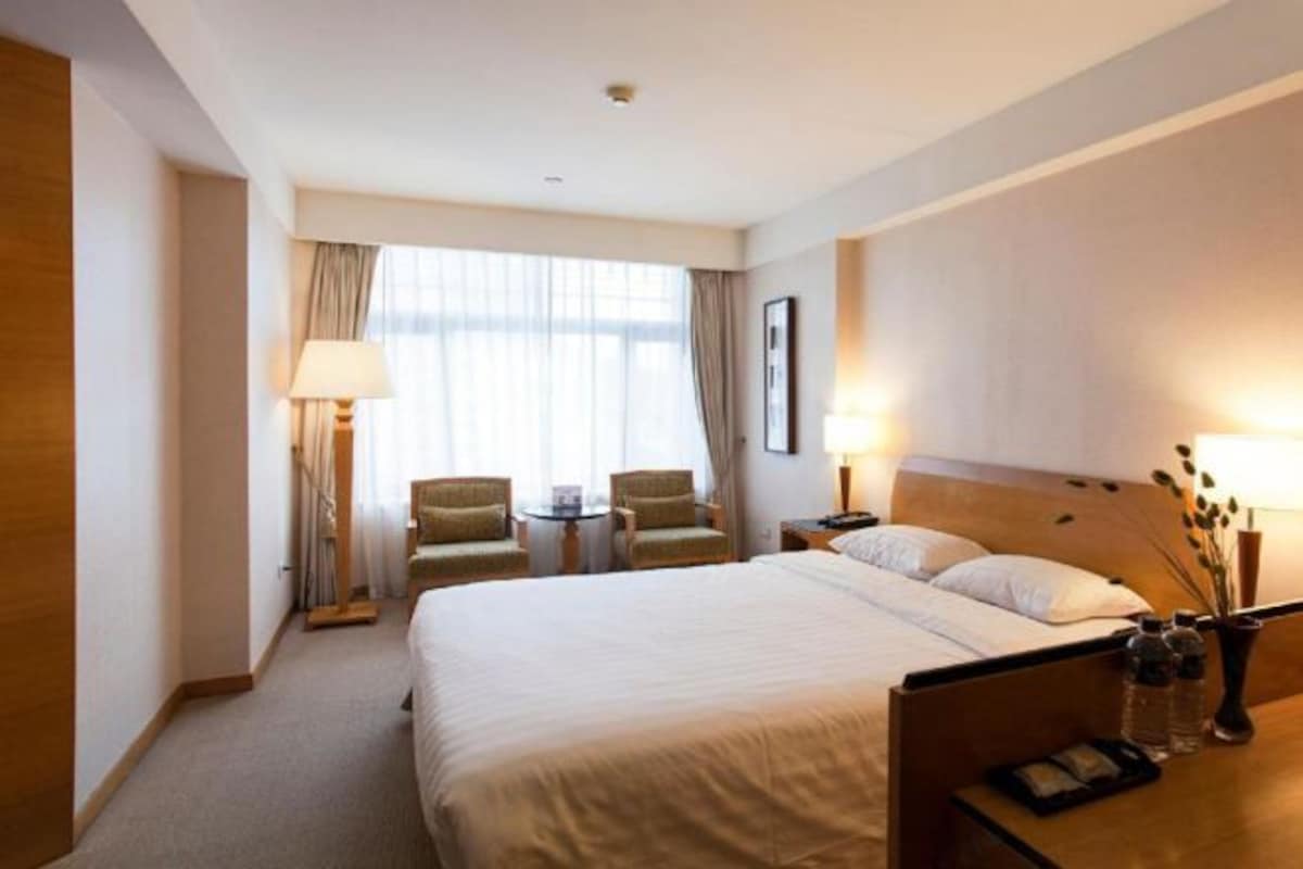 A hotel room with a large bed, two armchairs, a small table, and a nightstand with lamps and a vase.