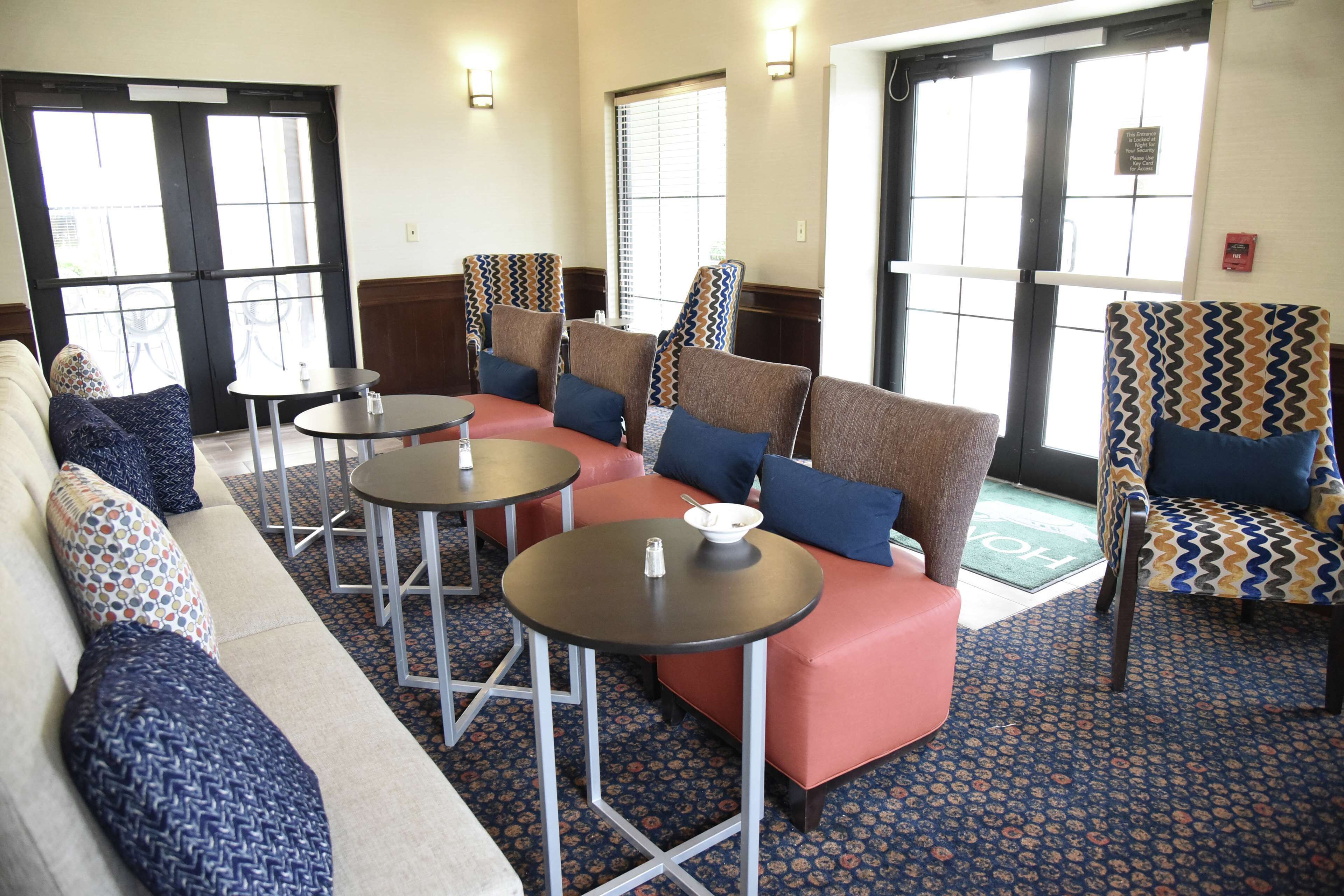 A hotel lobby with a sofa, chairs, and tables. There are windows with blinds, a fire extinguisher on the wall, and a patterned carpet.