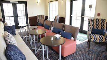 A hotel lobby with a sofa, chairs, and tables. There are windows with blinds, a fire extinguisher on the wall, and a patterned carpet.