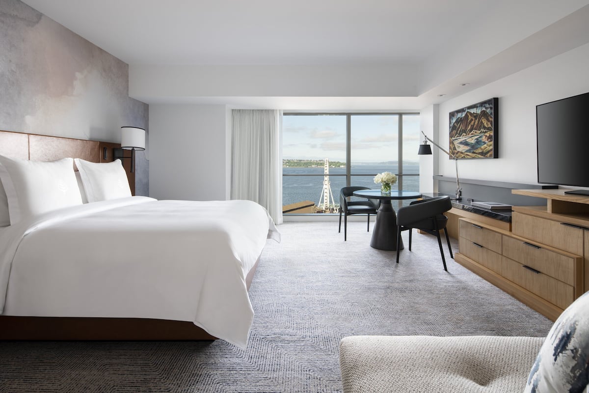A modern hotel room with a large bed, a desk with a chair, a television, and a view of the sea.