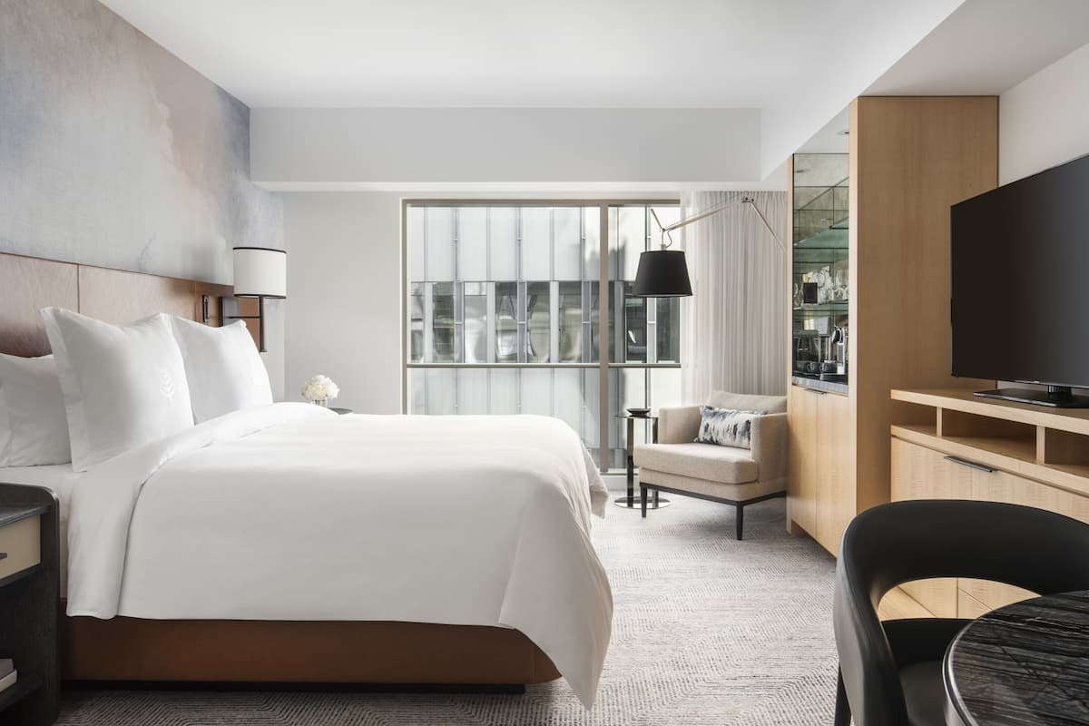 A modern hotel room with a large bed, a TV, a desk, and a view of the city through large windows.