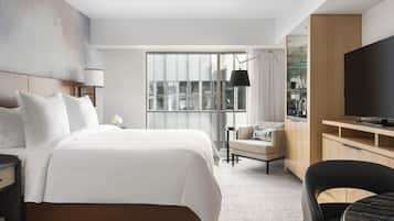 Habitación de hotel moderna con una cama grande, televisor, escritorio y vistas a la ciudad a través de amplias ventanas.
