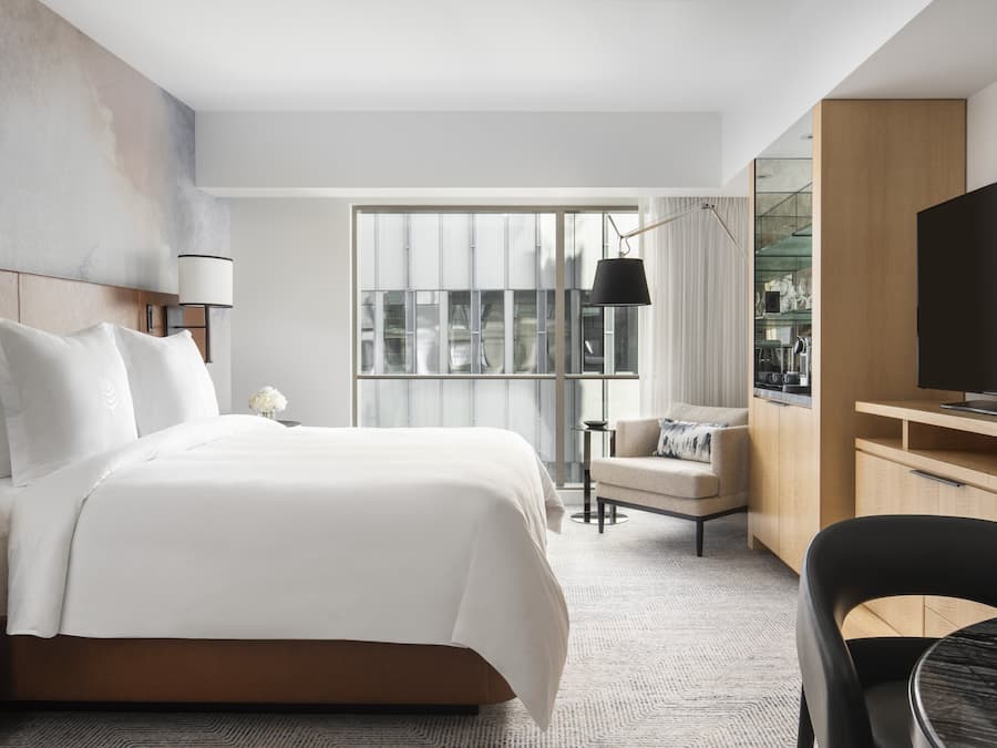 A modern hotel room with a large bed, a TV, a desk, and a view of the city through large windows.