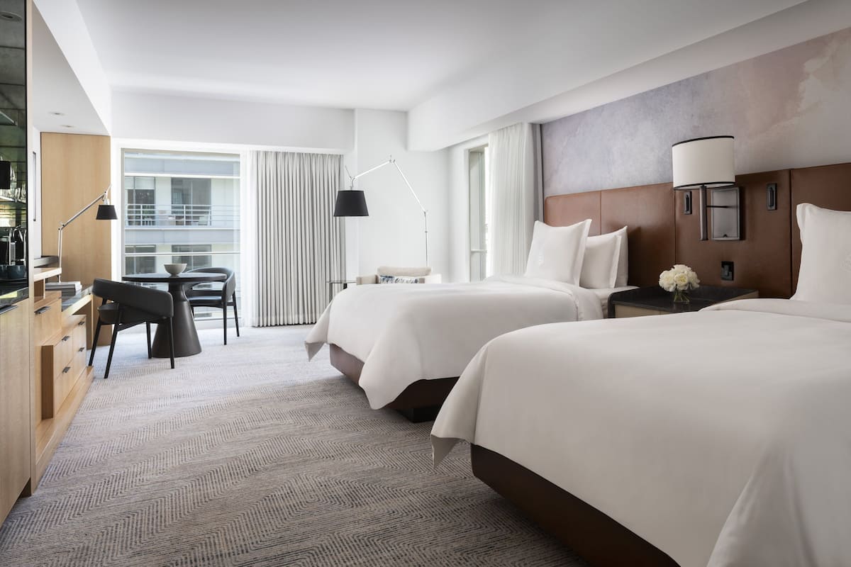 A hotel room with two beds, a desk, a chair, a lamp, and a view of the city.