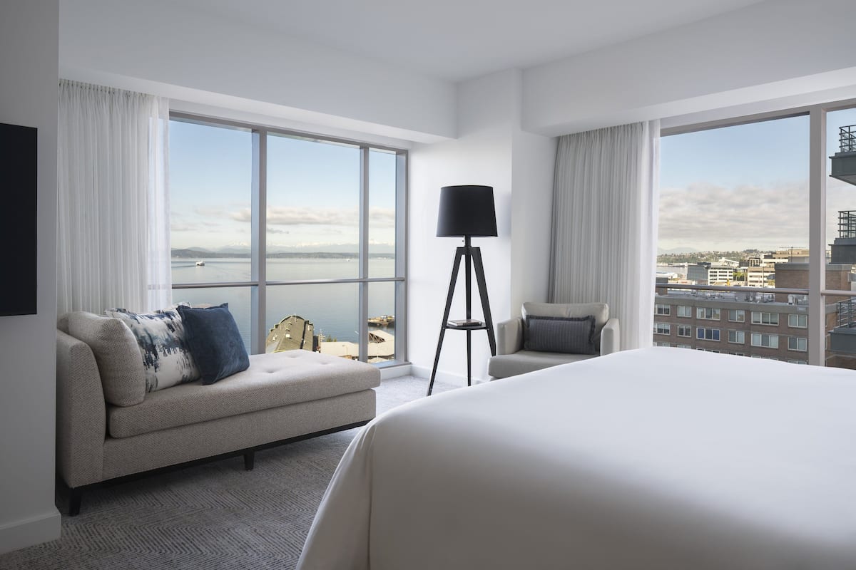 A modern hotel room with a large bed, a sofa, a chair, and a floor lamp. There are large windows offering a view of the city and a body of water.