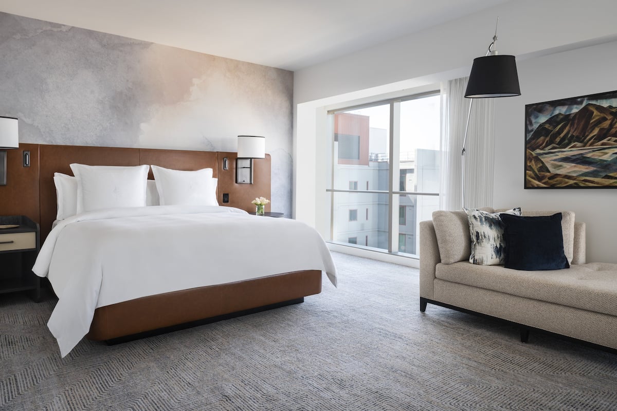 A modern hotel room with a large bed, a sofa, a large window with a view, and a painting on the wall.