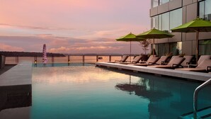 Un área junto a la piscina con sillones reclinables y sombrillas, con vista a una masa de agua y una noria a lo lejos.