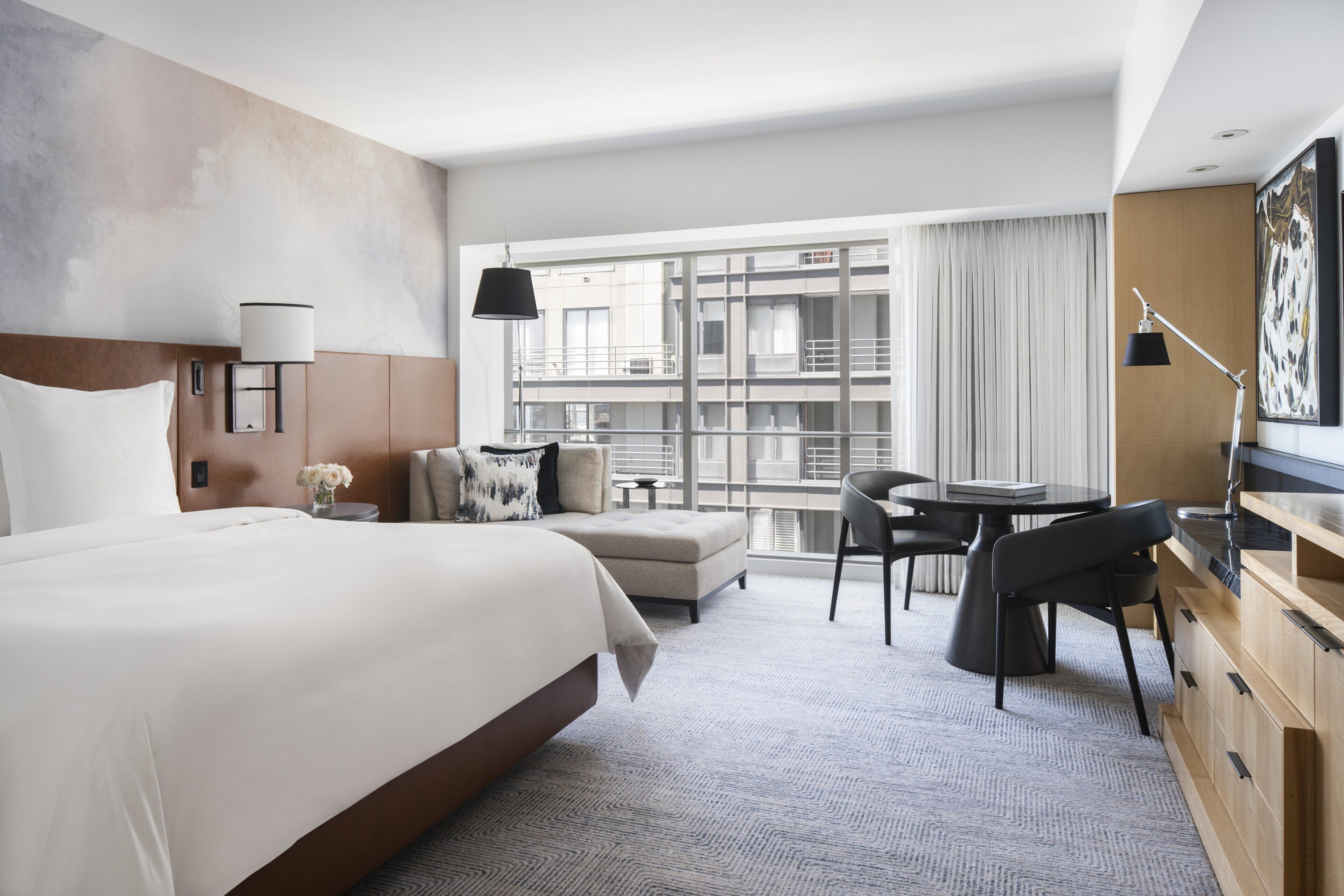 Ein modernes Hotelzimmer mit einem großen Bett, einem Schreibtisch, einem Sessel, einem kleinen Tisch und Blick auf die Stadt durch große Fenster.