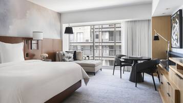 Une chambre d’hôtel moderne dotée d’un grand lit, d’un bureau, d’une chaise, d’une petite table et offrant une vue sur la ville grâce à de grandes fenêtres.