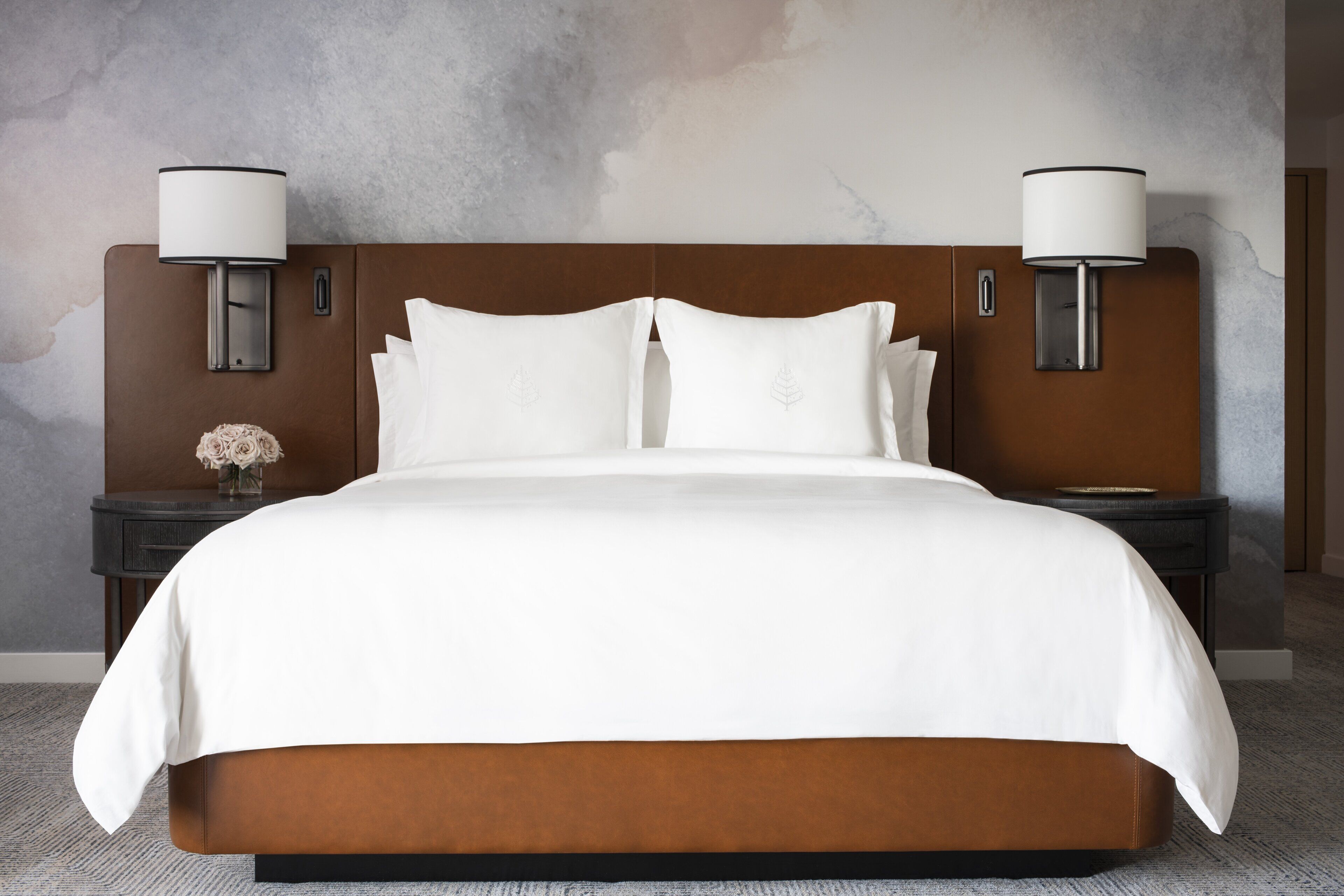 A large bed with a wooden headboard, two bedside tables with lamps, and a vase with flowers on one table.