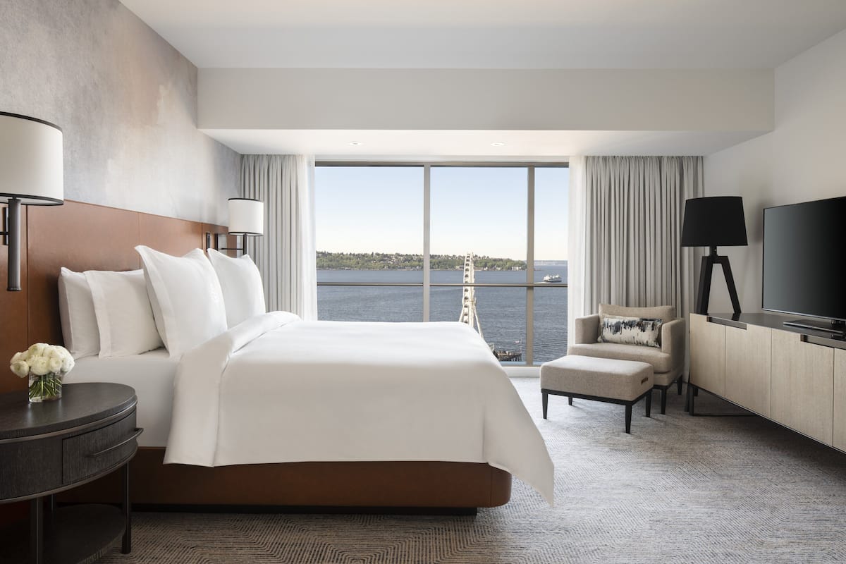 A modern hotel room with a large bed, a desk, a chair, a TV, and a view of the water.