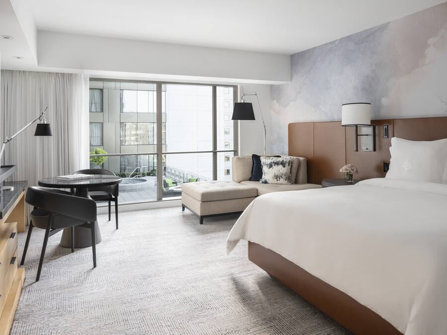 A modern hotel room with a large bed, a desk, a chair, a small table, and a balcony with a view.