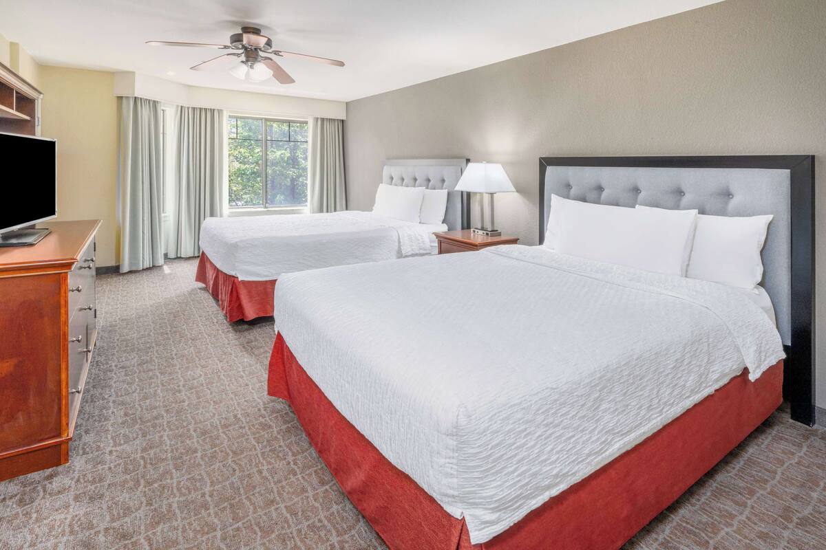 Une chambre d’hôtel avec deux lits, une télévision, un ventilateur de plafond et une grande fenêtre avec des rideaux.