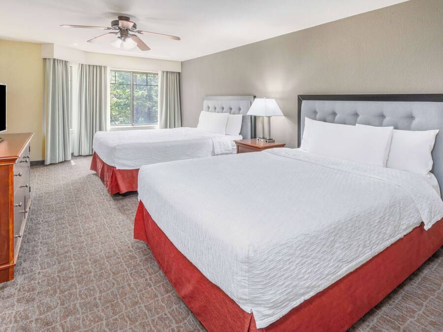 A hotel room with two beds, a TV, a ceiling fan, and a large window with curtains.