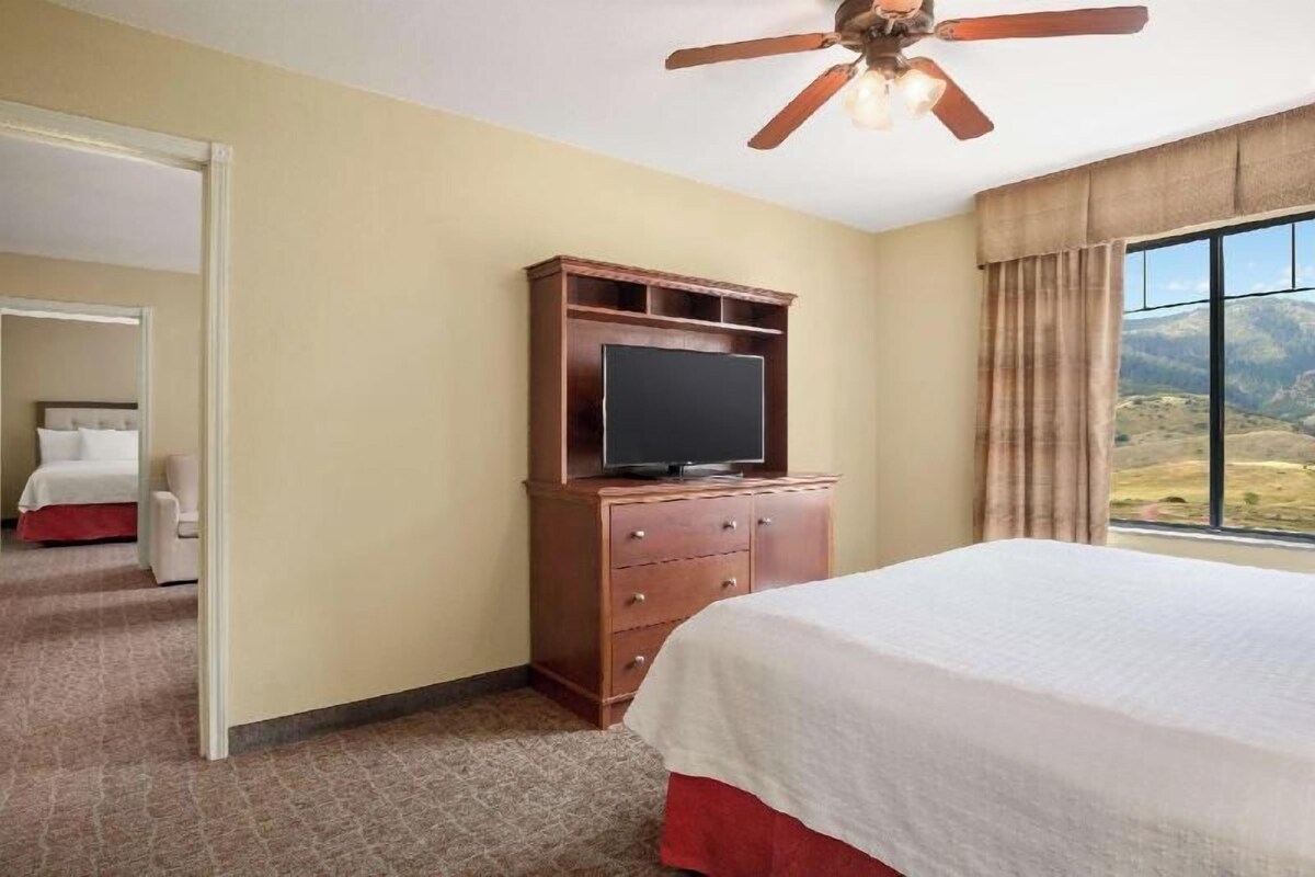 A hotel room with a bed, a TV on a wooden cabinet, a ceiling fan, and a window with a mountain view.