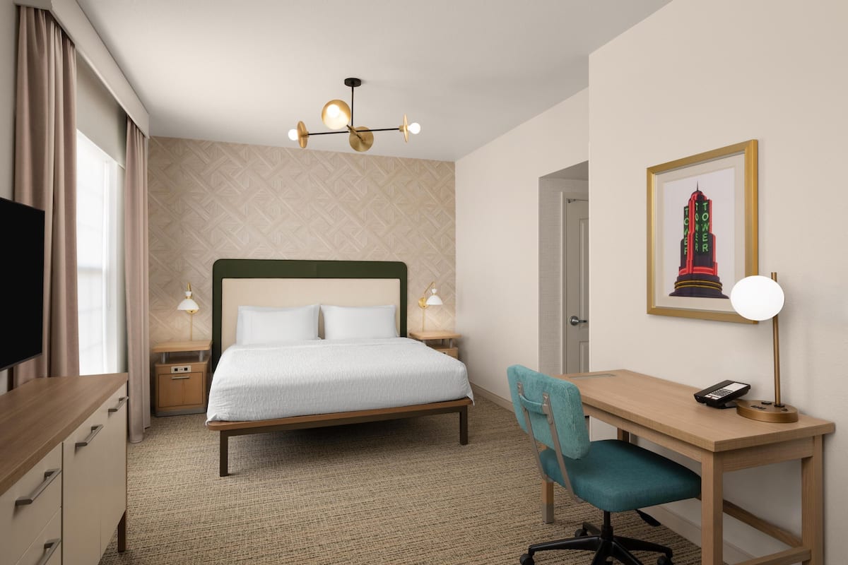 A hotel room with a bed, desk, chair, bedside lamps, a television, and a framed picture on the wall.