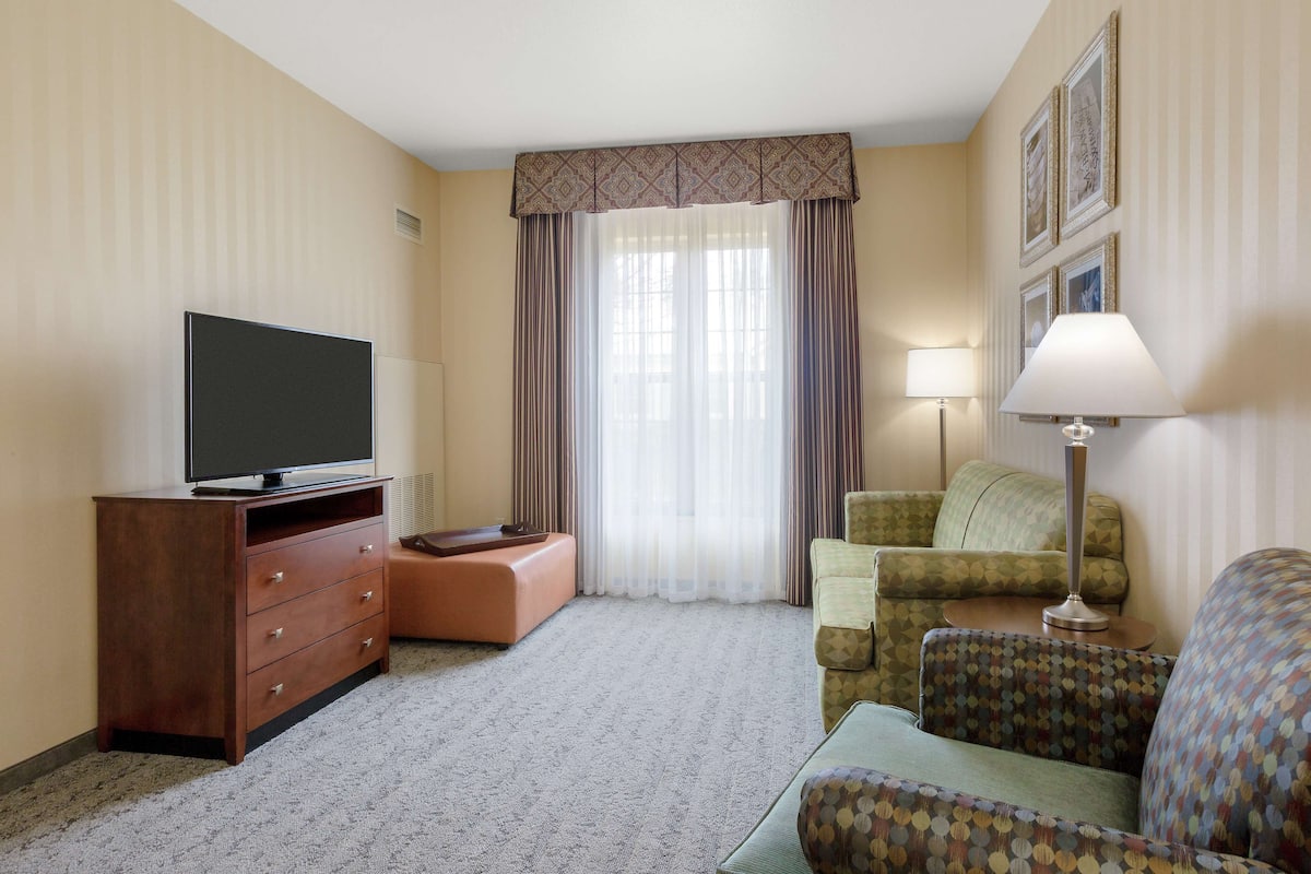 A hotel room with a flat-screen TV on a wooden cabinet, a green patterned sofa, a small round table with a lamp, and a large window with sheer curtains.