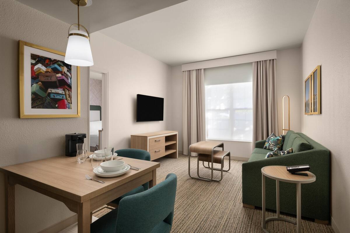 A hotel room with a dining table, chairs, a sofa, a TV, and a window with curtains.