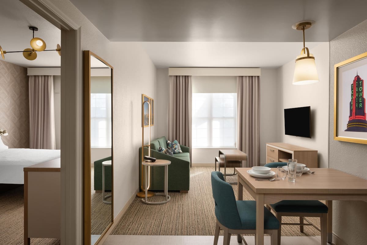 A hotel room with a bed, a green sofa, a dining table with chairs, a small round table with a lamp, and a TV on the wall.