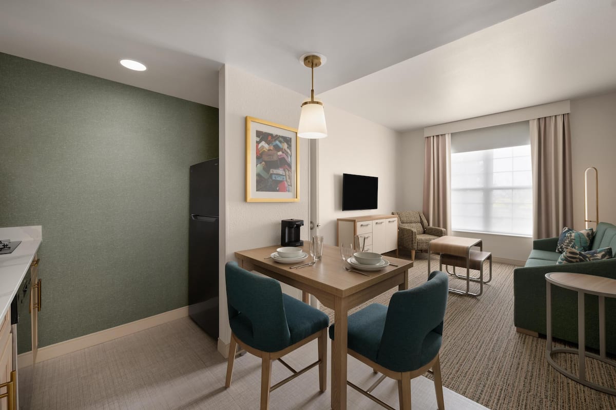 A hotel room with a dining area, a sofa, a TV, and a window with curtains.