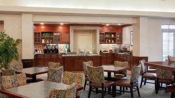 A hotel dining area with multiple tables and chairs, a kitchen in the background, and a large potted plant.