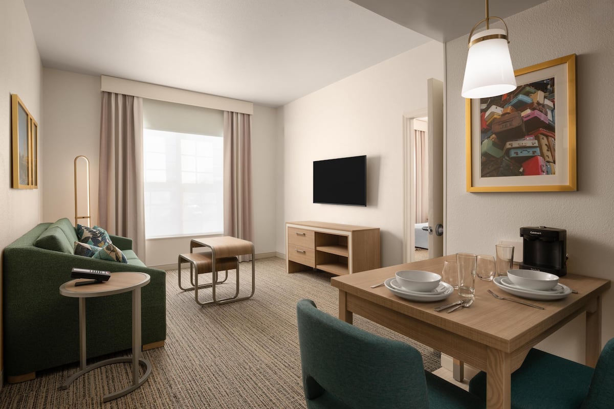 A hotel room with a green sofa, a small round table, a wooden desk, a television, and a dining area with chairs and plates.