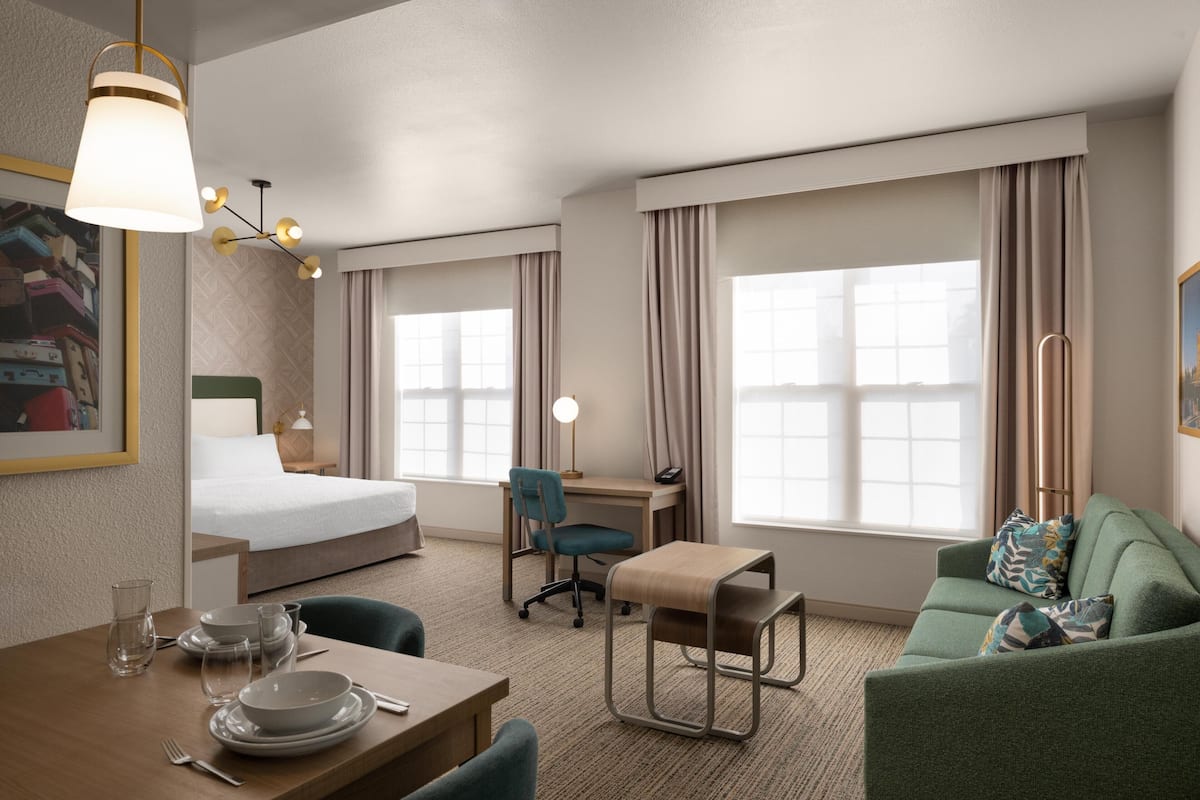 A hotel room with a bed, a desk, a green sofa, a dining table with plates and glasses, and large windows with curtains.