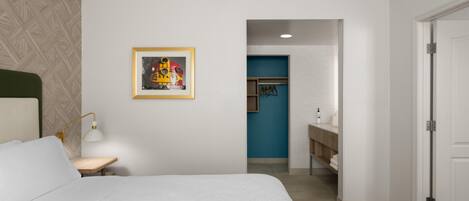 A hotel room with a bed, a desk, a chair, a lamp, a framed picture on the wall, and a bathroom visible through an open door.
