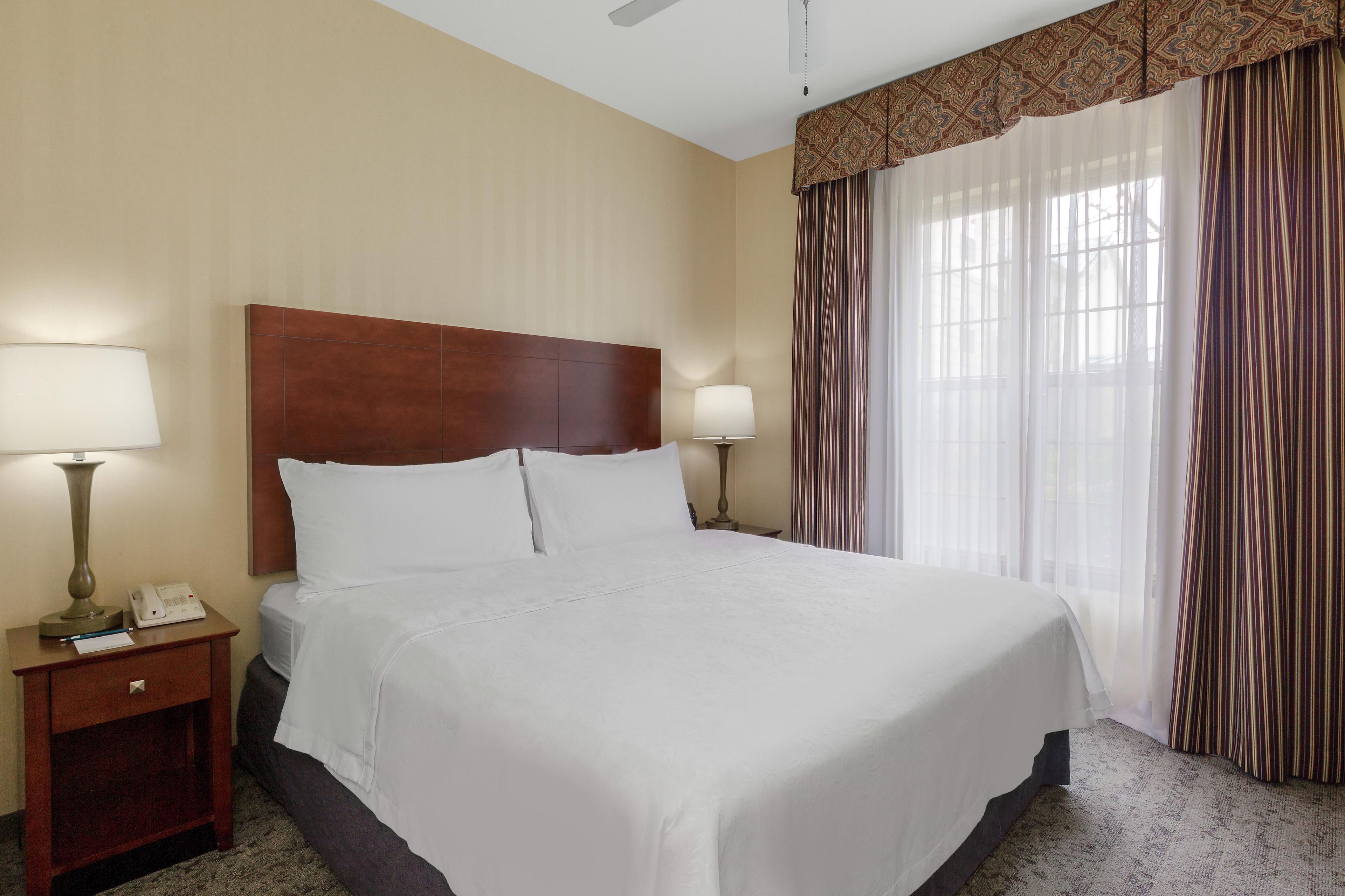 Una habitación de hotel con una cama grande, dos lámparas de noche, una mesita de noche y un ventanal con cortinas.