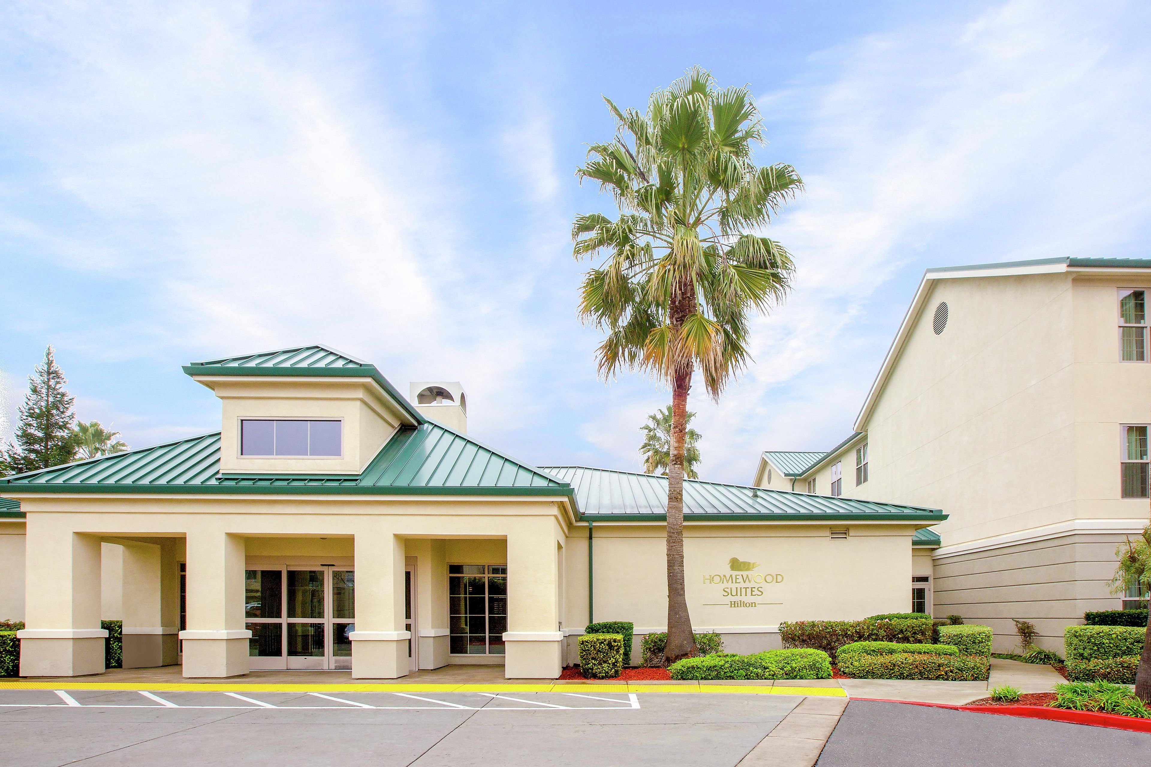 A hotel with a green roof, a palm tree in front, and a sign that reads 'HOMEWOOD SUITES Hilton'.