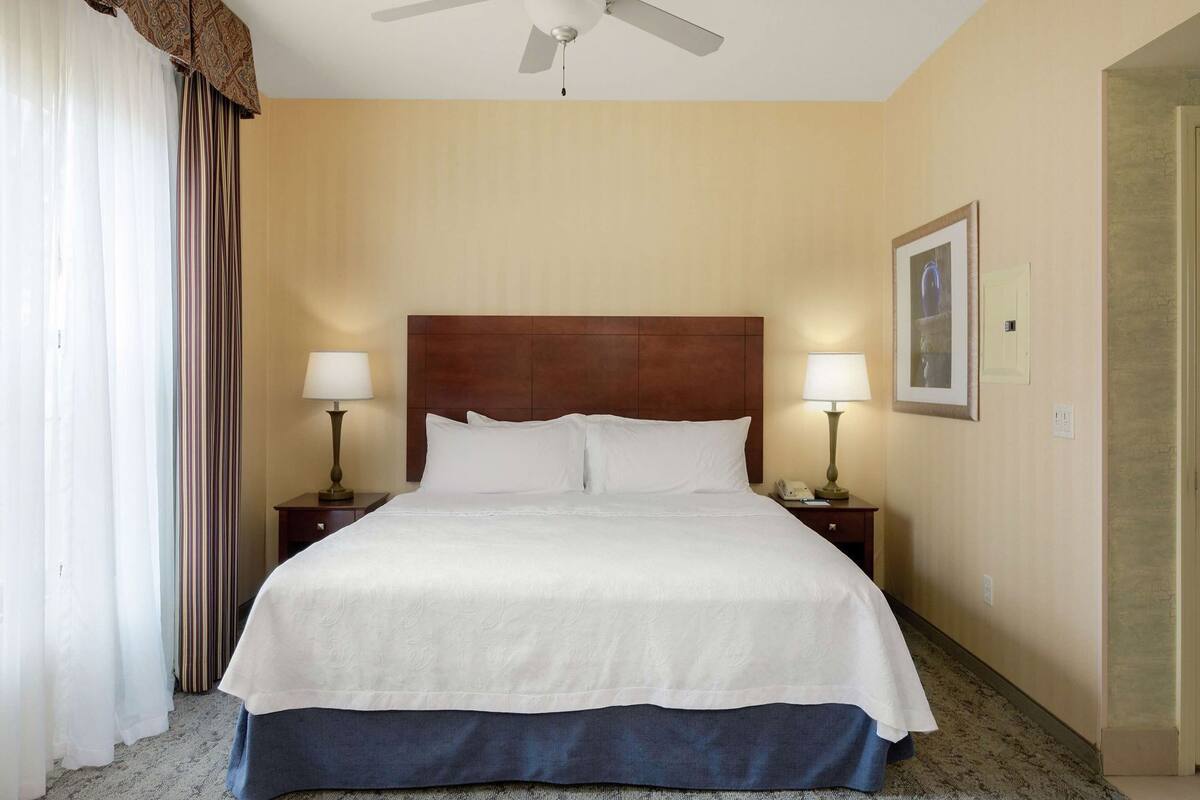 A hotel room with a large bed, two bedside tables with lamps, a ceiling fan, and a framed picture on the wall.