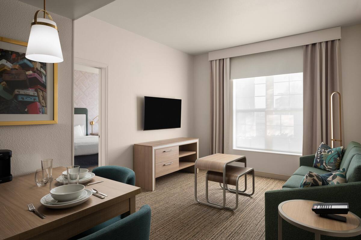 A hotel room with a dining area, a sofa, a TV, and a large window with curtains.