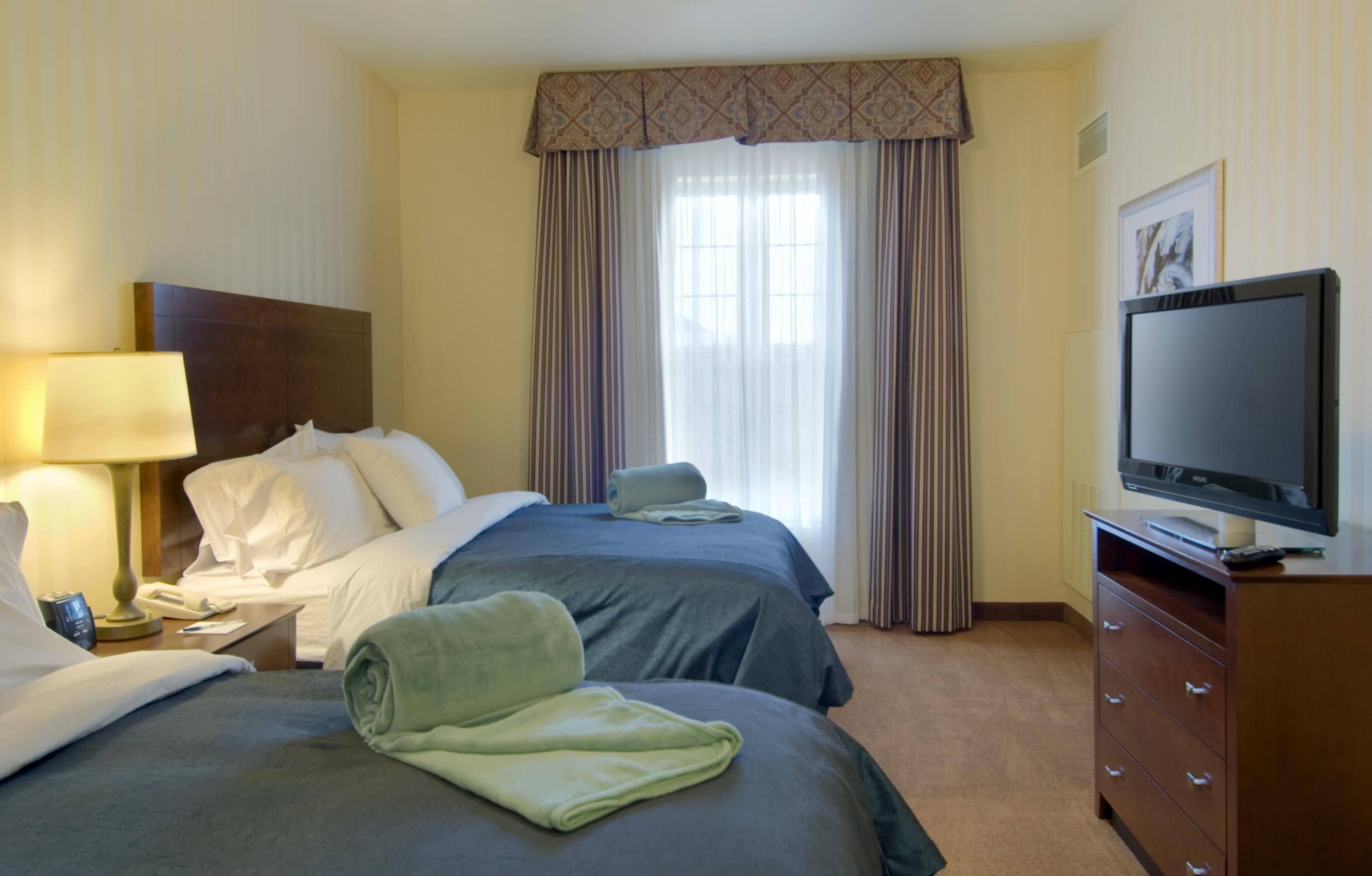 A hotel room with two beds, a television, a desk with a lamp, and a window with curtains.