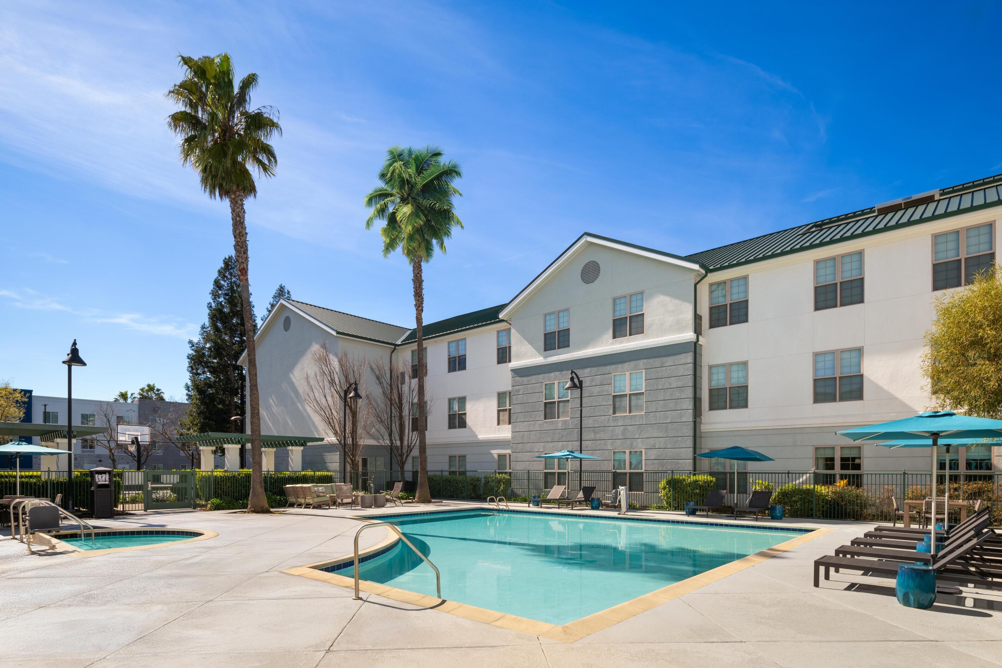 A pool area with lounge chairs and umbrellas, a building with multiple windows, and palm trees.