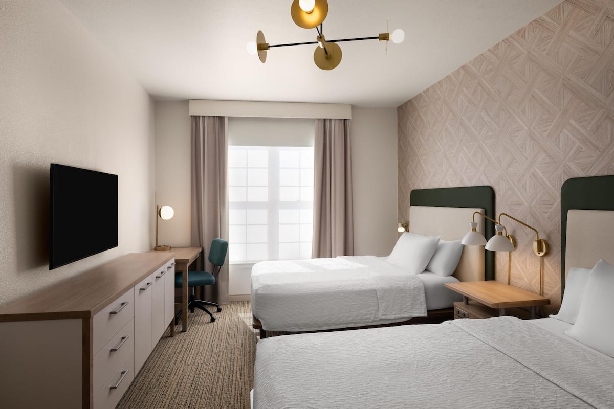 A hotel room with two beds, a desk, a chair, a TV, and a window with curtains.