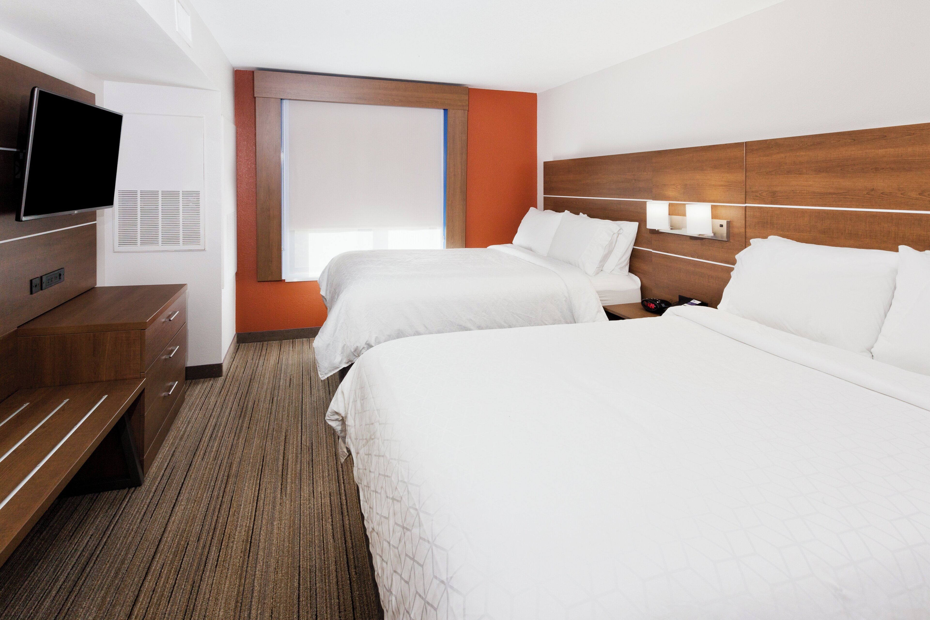 A hotel room with two beds, a TV, a desk, and a window with blinds.