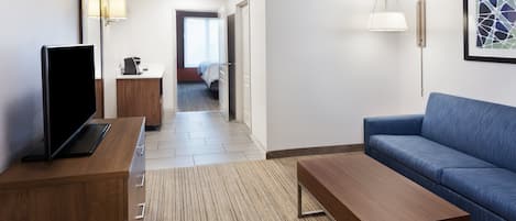 A hotel room with a wooden desk, a blue sofa, a flat-screen TV, a bedside table, a lamp, and a framed artwork on the wall.