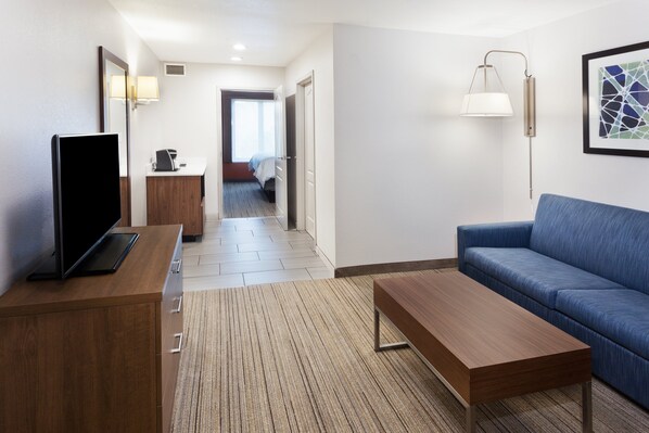 A hotel room with a wooden desk, a blue sofa, a flat-screen TV, a bedside table, a lamp, and a framed artwork on the wall. - Holiday Inn Express & Suites Rome-East by IHG (Rome)