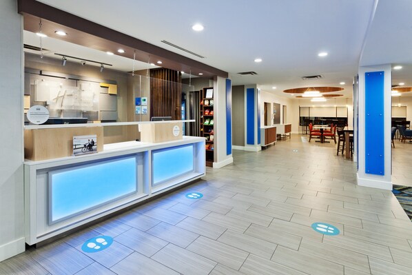 Uma área de recepção de hotel com design moderno, incluindo um balcão de recepção, sala de estar e um espaço de refeições visível ao fundo.