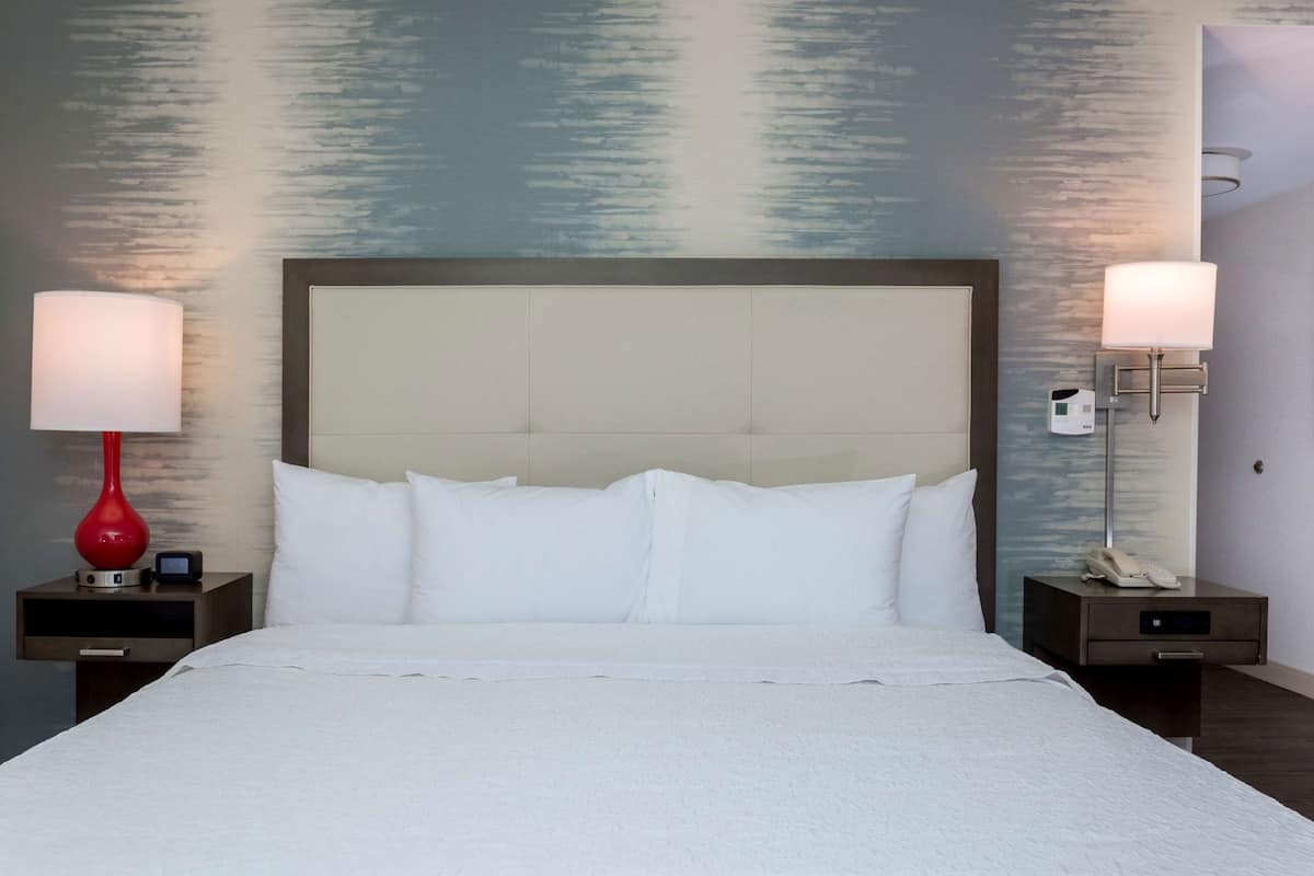 A hotel room with a large bed, two bedside tables, a red lamp, and a wall-mounted phone.