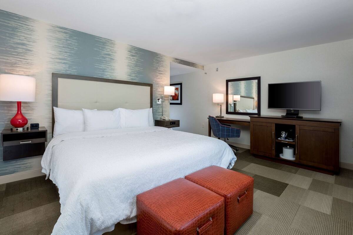 A hotel room with a large bed, a desk with a chair, a television, and a small red suitcase.